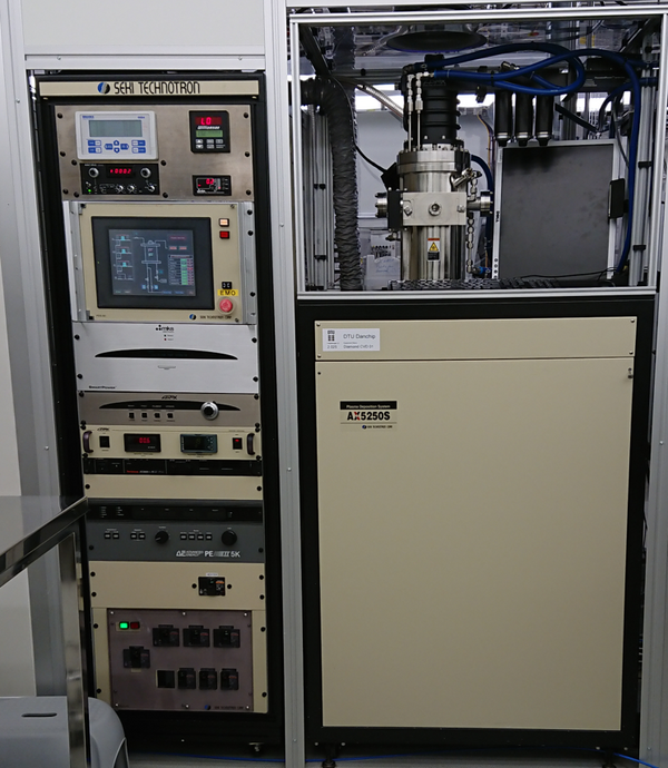 On the left is a tall instrument rack with various panels and control screens. It says "Seki technotron" on top. There are indicator lamps and an EMO button. On the right is the processing chamber which is relatively small, it's a cylindrical vacuum chamber with a small window facing the viewer. It's connected to gas lines and an exhaust system on top. There's a large panel covering the lower part of the instrument.